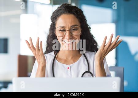 Photo en gros plan d'une jeune femme hispanique médecin parlant au patient et souriant, utilisant un ordinateur portable pour les appels vidéo, consultant regardant l'écran d'un ordinateur portable, travaillant dans le bureau de la clinique. Banque D'Images