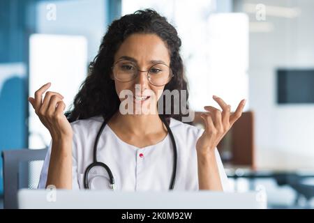 Photo en gros plan d'une jeune femme hispanique médecin parlant au patient et souriant, utilisant un ordinateur portable pour les appels vidéo, consultant regardant l'écran d'un ordinateur portable, travaillant dans le bureau de la clinique. Banque D'Images