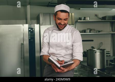 Un chef souriant en uniforme prend des notes dans un carnet situé dans la cuisine Banque D'Images