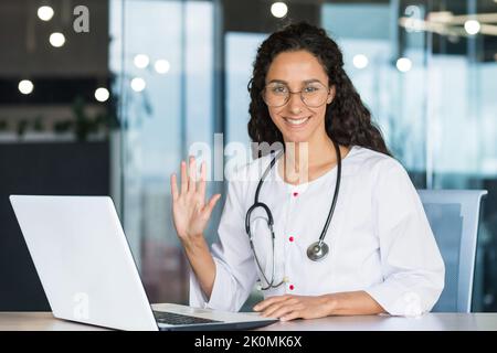 Portrait d'une jeune femme médecin professionnelle d'Amérique latine sous un manteau médical blanc souriant et regardant l'appareil photo tenant sa main vers le haut geste de salutation, la femme médecin travaille à l'intérieur Banque D'Images