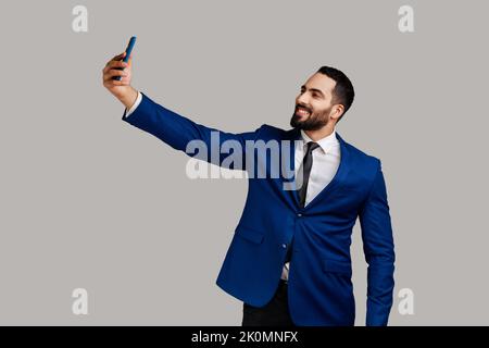 Homme barbu qui fait le selfie sur un appareil photo de smartphone, blogueur communiquant, enregistrant des vidéos pour les abonnés des réseaux sociaux, portant un costume officiel. Prise de vue en studio isolée sur fond gris. Banque D'Images