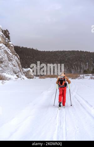 Vue arrière d'un jeune homme en vêtements brun rouge avec sac à dos ski près des rochers et des falaises Active sain style de vie sports d'hiver randonnée pédestre Banque D'Images