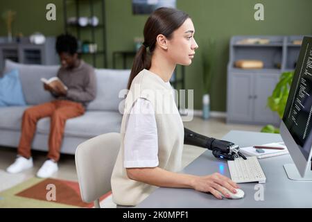 Jeune fille sérieuse avec une incapacité travaillant avec des codes sur ordinateur à son lieu de travail à la maison avec l'homme lisant le livre en arrière-plan Banque D'Images