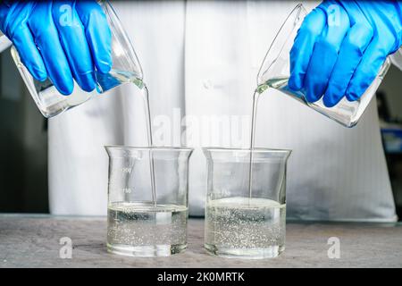 Une personne dans une blouse de laboratoire verse deux liquides transparents dans les béchers Banque D'Images