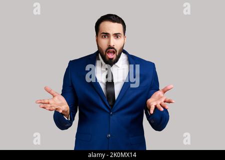 Portrait d'un homme barbu debout avec des mains levées et expression indignée, demandant la raison du conflit, portant un costume officiel. Prise de vue en studio isolée sur fond gris. Banque D'Images