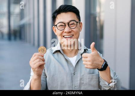 Un homme d'affaires asiatique heureux montrant le bitcoin et faisant des gestes près d'un immeuble de bureaux moderne, à l'extérieur Banque D'Images