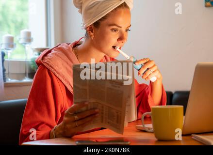 Image symbolique Precrastination, la jeune femme tente de faire beaucoup de choses en même temps, après le bain, sur l'ordinateur, téléphone, journal, et le petit déjeuner Banque D'Images