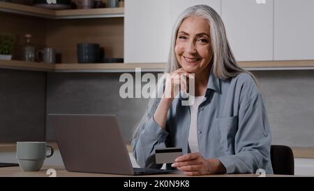 Vieille dame acheteur grand-mère cashopper femme mature assis dans la cuisine payer les factures d'utilité sur ordinateur portable utiliser l'application bancaire pour la charité en ligne entrer la carte de sécurité Banque D'Images