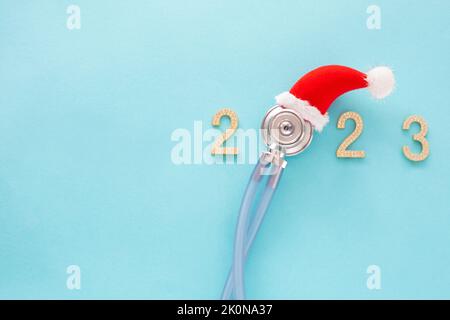 Stéthoscope médical dans un chapeau de Père Noël rouge et les chiffres 2023 sur fond bleu. Concept Noël et nouvel an. Hiver médical créatif Banque D'Images