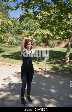 Fitness jeune femme redhead faisant de l'exercice d'étirement à l'extérieur dans le parc public. Banque D'Images