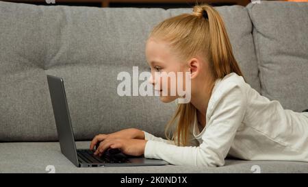 Insouciant petit enfant jolie fille blonde enfant fille utilisant un ordinateur portable couché sur un canapé gris surfer sur Internet chat avec des camarades de classe jouant des jeux d'étude Banque D'Images
