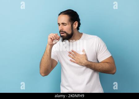 Portrait d'un malade malsain avec une barbe et des cheveux recueillis portant un T-shirt blanc ayant des symptômes de grippe, toux, gardant la main près de la bouche. Studio d'intérieur isolé sur fond bleu. Banque D'Images