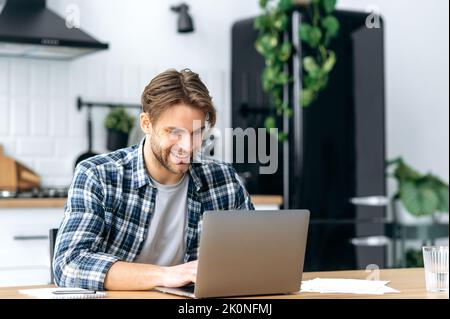 Satisfait caucasien positif jeune homme, élégamment habillé, indépendant, SPÉCIALISTE INFORMATIQUE, crypto Trader, s'assoit dans la cuisine à la maison, travaillant à distance en ligne sur un nouveau projet, tapant sur un clavier, sourit Banque D'Images