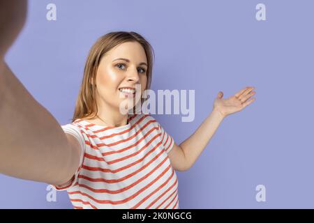 Portrait d'une femme blonde sympathique positive portant un T-shirt rayé montrant le geste de bienvenue, invitant à sa maison, faisant selfie tourné pov sur le téléphone mobile. Studio d'intérieur isolé sur fond violet Banque D'Images