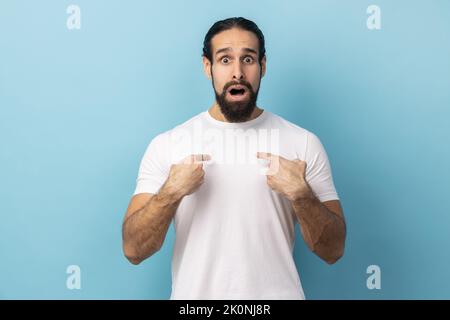 Portrait d'un homme heureux et impressionné avec une barbe portant un T-shirt blanc pointant vers lui, se sentant fier et ambitieux, choqué par un énorme succès. Studio d'intérieur isolé sur fond bleu. Banque D'Images