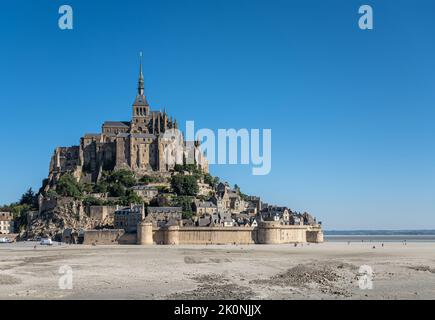 Mont Saint-Michel, Normandie, France - 8 juillet 2022 : la structure en pierre brune de sa roche entourée d'appartements sablonneux à marée basse contre ciel bleu. Banque D'Images