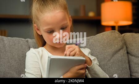 Smart concentrée écolière enfant caucasien école primaire élève focalisé écriture essai faire des devoirs de maths assis sur le canapé faire des notes dans le carnet Banque D'Images