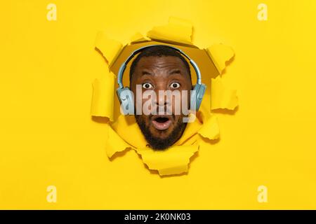 Homme noir émotif écoutant de la musique préférée via un casque et regardant l'appareil photo à travers le trou de fond jaune Banque D'Images