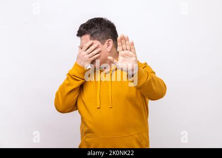 Portrait d'un homme confus couvrant les yeux et levant la main pour s'arrêter, se sentant stressé peur, refusant de regarder le contenu effrayant, portant le sweat à capuche de style urbain. Studio d'intérieur isolé sur fond blanc. Banque D'Images