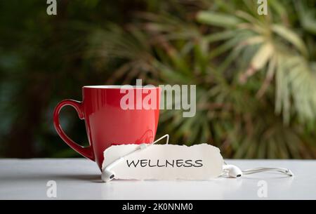 Word, Wellness sur un morceau de papier blanc à côté d'une tasse rouge avec des écouteurs. Concept d'auto-soins et de bien-être. Banque D'Images