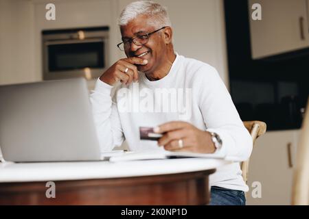 Un homme de grande garde souriant gaiement tout en faisant des achats en ligne à la maison. Heureux homme mature utilisant sa carte de crédit pour passer une commande en ligne sur un ordinateur portable. Stylo Banque D'Images