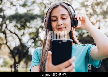 Une adolescente porte un t-shirt turquoise, utilise un casque et établit une liste de lecture musicale sur son smartphone dans le parc Banque D'Images