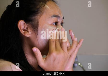 Femme asiatique se laver le visage dans la salle de bains après la douche. Femme heureux nettoie la peau avec de la mousse dans la salle de bains. Banque D'Images