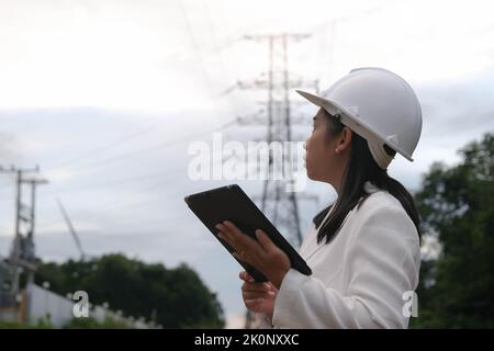 Femme asiatique électricien travaillant sur une tablette près d'un pôle haute tension, inspectant le réseau électrique. Ingénieur en éoliennes pour la production d'énergie électrique. Banque D'Images