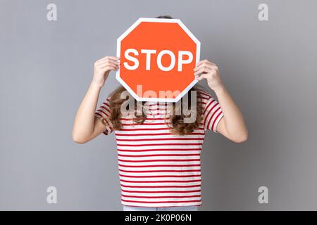 Petite fille en T-shirt rayé couvrant le visage avec le symbole Stop, femme anonyme tenant le feu rouge stop, avertissement de danger, restriction et limites. Prise de vue en studio isolée sur fond gris. Banque D'Images