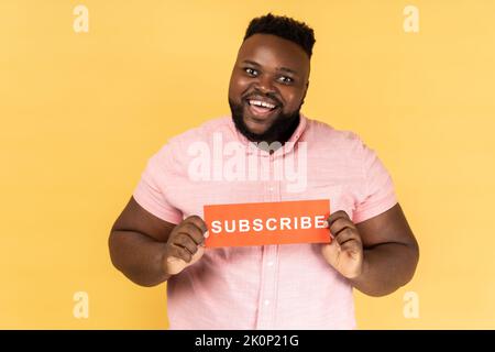 Portrait d'homme barbu positif gai portant une chemise rose tenant une carte rouge avec inscription de souscription, demandant de suivre son vlog. Studio d'intérieur isolé sur fond jaune. Banque D'Images