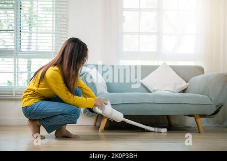 Femme de ménage nettoyage de la poussière sol sous canapé ou mobilier de canapé avec aspirateur, jeune femme asiatique heureuse avec accumulateur aspirateur à hom Banque D'Images