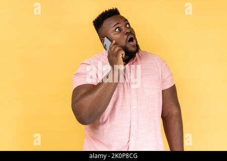 Portrait d'un homme choqué portant une chemise rose discutant avec un ami sur un téléphone portable, regardant loin avec surpris étonné, entendre des nouvelles de rupture. Studio d'intérieur isolé sur fond jaune. Banque D'Images