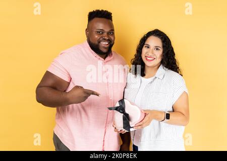 Portrait d'un jeune couple souriant ravie en vêtements décontractés debout ensemble, homme pointant sur la boîte actuelle entre les mains de sa femme, célébrant les vacances. Studio d'intérieur isolé sur fond jaune. Banque D'Images