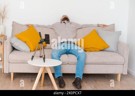 Homme barbu souriant filmant de la vidéo en utilisant un smartphone sur un trépied en ligne tout en étant assis sur un canapé dans le salon, portant une chemise et un Jean. Influence des médias sociaux, création de contenu et consultation en ligne Banque D'Images