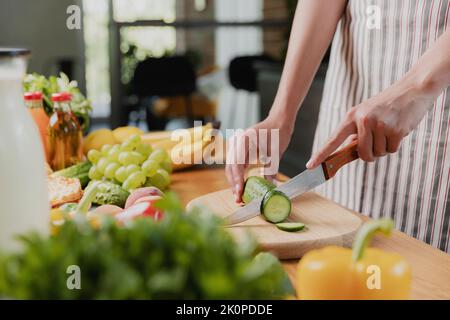 Gros plan sur la jeune femme de ménage, les mains de fille tranchant le concombre avec un couteau de cuisine, coupe les aliments sur une planche à découper en bois. Femme méconnue au comptoir de cuisine, coupant des légumes biologiques Banque D'Images