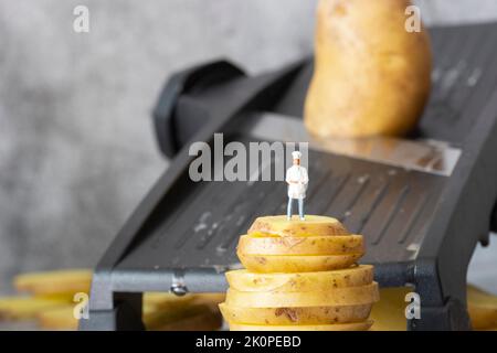 Petit chef modèle échelle, debout sur des pommes de terre empilées, avec coupe-cuisine mandoline en arrière-plan Banque D'Images