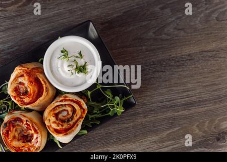 Appétissants petits pains de pâte pepperoni avec garniture, verdure et sauce sur une élégante assiette noire sur une table en bois au restaurant Banque D'Images