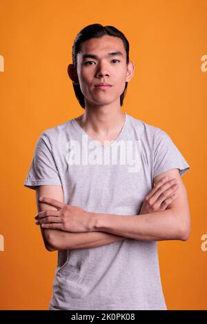 Jeune homme asiatique sérieux debout dans une posture confiante avec les bras croisés, regardant la caméra, vue de face. Portrait réussi de l'adolescent avec expression du visage détendue, prise de vue moyenne Banque D'Images