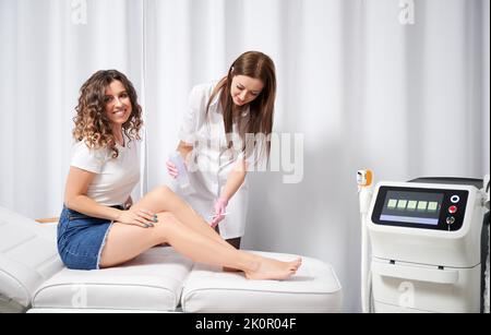 Portrait de la jeune femme heureuse que l'esthéticienne appliquant le gel à la jambe avant la procédure d'enlèvement de cheveux au laser des cheveux indésirables. Concept de la procédure cosmétique confortable populaire pour l'enlèvement de cheveux de laser. Banque D'Images