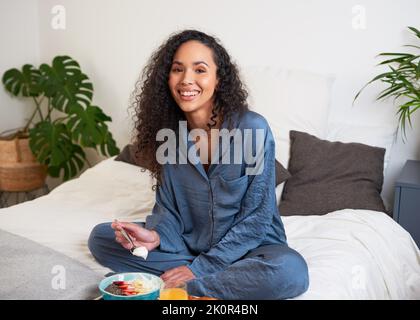 Une belle jeune femme mange le petit déjeuner au lit en pyjama de soie le week-end Banque D'Images
