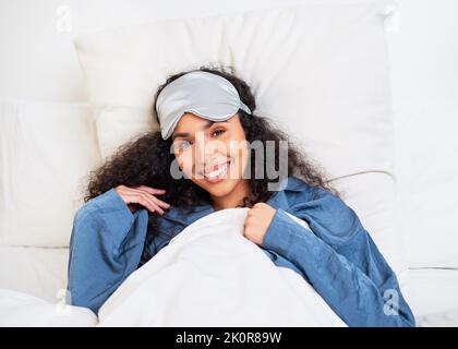 Une jeune femme multiethnique s'étire avec les bras derrière la tête et le masque de sommeil Banque D'Images