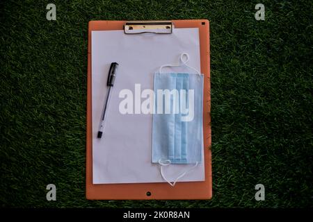 Vue rapprochée en haut d'un presse-papiers en bois avec un papier, un stylo noir et un masque sur de l'herbe verte Banque D'Images