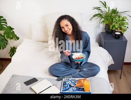 Une belle jeune femme mange le petit déjeuner au lit en pyjama de soie le week-end Banque D'Images