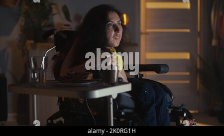 Femme handicapée assise en fauteuil roulant motorisé devant la télévision et se détendant à la maison la nuit en regardant un spectacle télévisé Banque D'Images