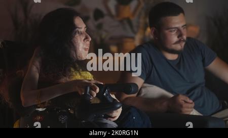 Homme aidant la femme avec l'atrophie musculaire spinale dans un fauteuil roulant pour utiliser la manette de jeu et ils jouent au jeu vidéo tout en passant la soirée ensemble à la maison Banque D'Images