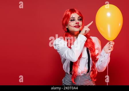 une femme heureuse avec des cheveux brillants et un maquillage clown pointant vers le ballon jaune isolé sur le rouge Banque D'Images
