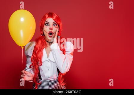 femme à tête rouge émerveillement dans le maquillage clown tenant le ballon jaune et touchant le visage isolé sur le rouge Banque D'Images