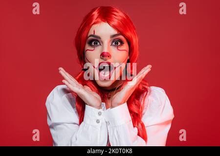 femme à tête rouge émerveillement avec maquillage d'halloween tenant les mains près du visage isolé sur le rouge Banque D'Images
