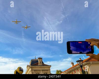 Un vol en formation de deux avions légers ouvre le départ du 36e Marathon des Châteaux du Médoc à Pauillac, en France, et tous les regards se tournent vers le ciel. Un spectateur capture la démonstration sur un smartphone, deux traînées de condensation croisent le ciel bleu, les toits de la ville sont visibles en dessous. Banque D'Images