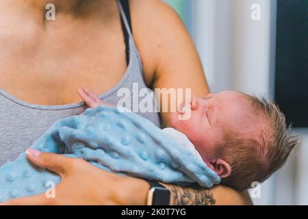 Adorable bébé garçon de nouveau-né enveloppé dans une couverture dormant paisiblement dans les bras de sa mère à la maison. Photo de haute qualité Banque D'Images